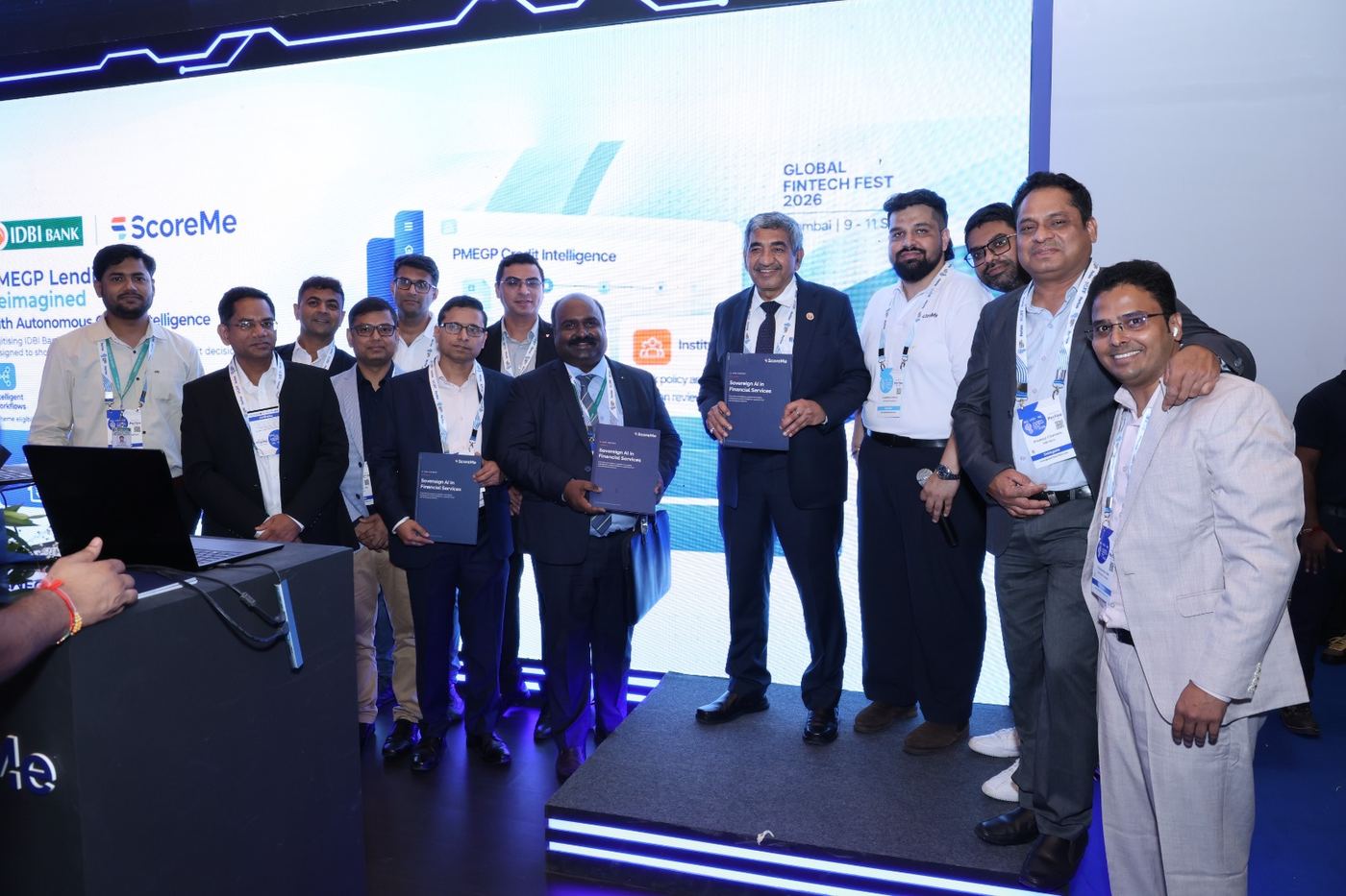 A group on stage holding copies of the report in front of an IDBI Bank and ScoreMe screen.